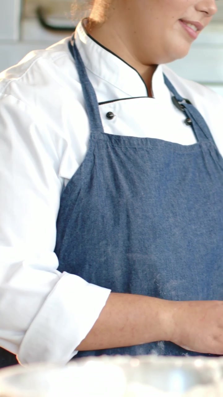 Video verticale: Chef che si china in avanti, con camice bianco e grembiule blu, che si allunga e prepara sul bancone.