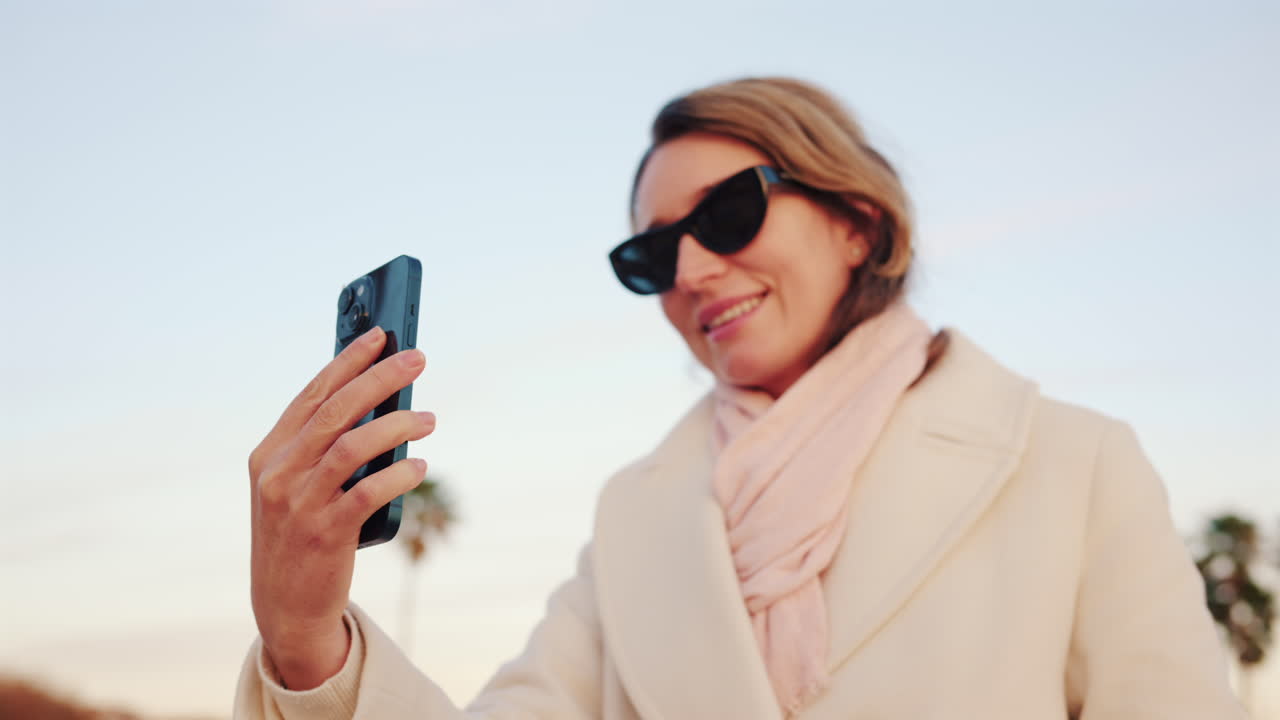 Donna sicura di sé con un camice bianco che scatta un selfie con il suo smartphone in una zona esterna soleggiata, fiancheggiata da palme