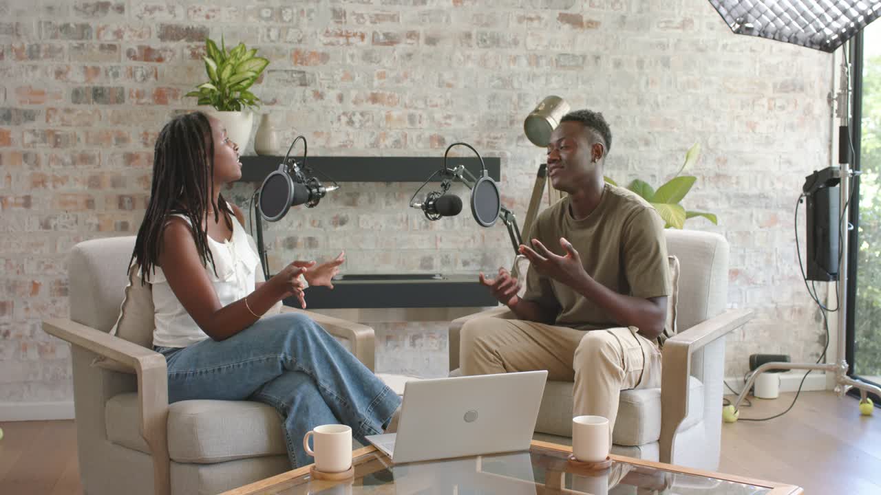African American co-hosts sitting in studio by brick wall, man initiating podcast using mics