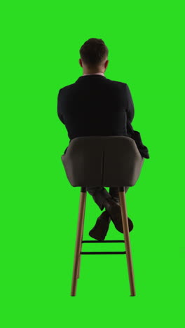 vertical video rear view studio shot of man sitting on stool facing away from camera against green screen 1