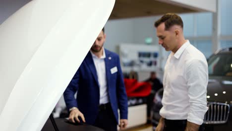 professional salesman showing car engine under open hood to couple in auto showroom