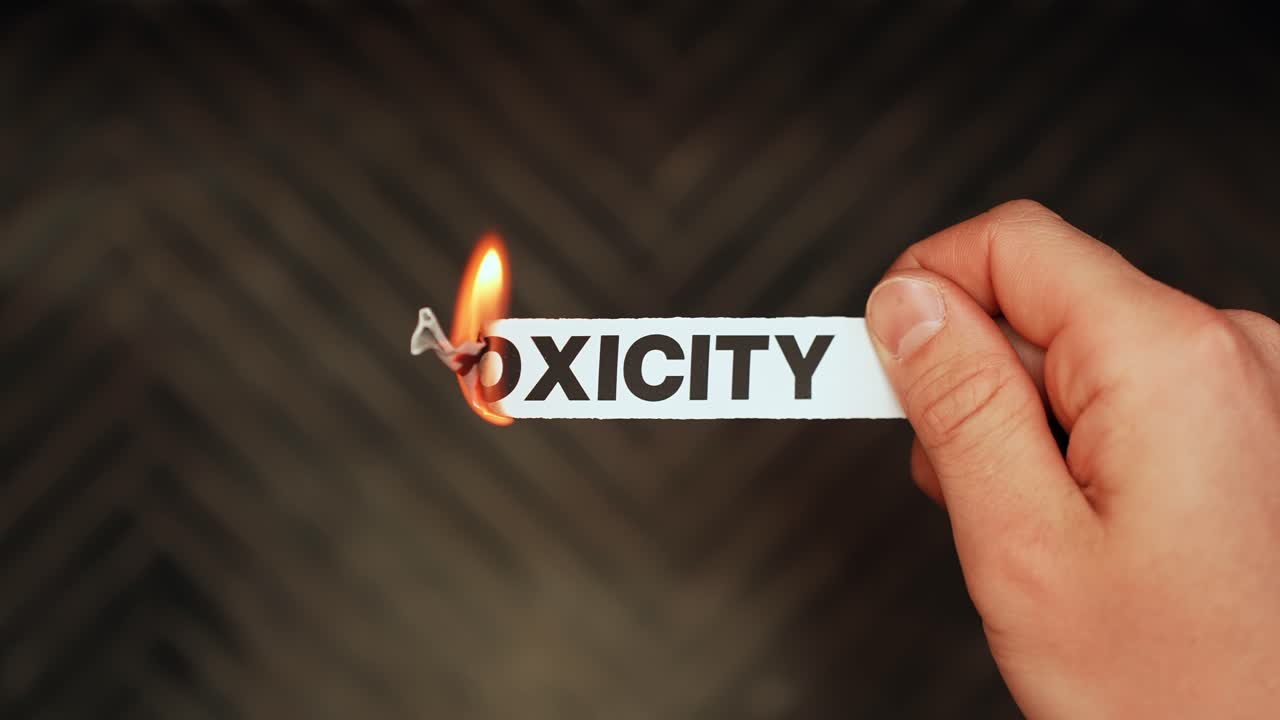 Close-up of a hand holding a paper strip labeled “TOXICITY” as the edge burns with flame on a dark background. Cutting toxic habits and cleansing negative energy concept