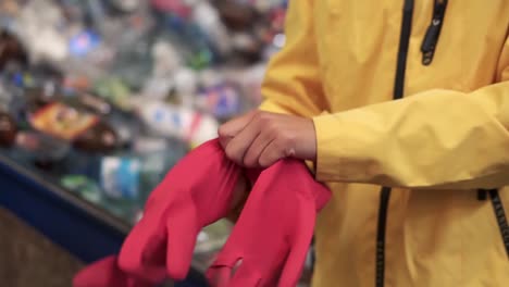 Close-up-view-of-woman-standing-indoor-put-red-rubber-protective-gloves-on-hands.-Caution-preventive-measures-working-with-dirt.-Self-care,-personal-safety.-Pile-of-used-plastic-bottles-on-blurred-background