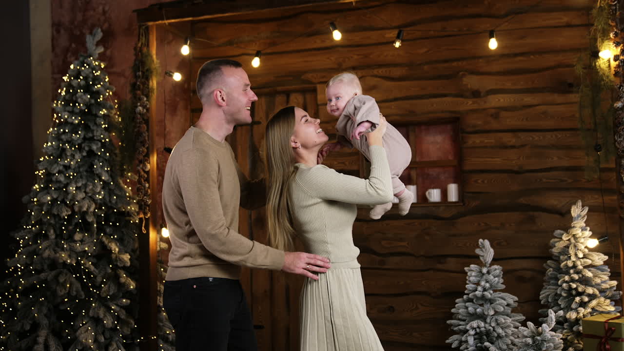 Smiling woman holding a cute little blond infant over her head. Man stands behind his wife holding her by the waist. Christmas trees at backdrop.