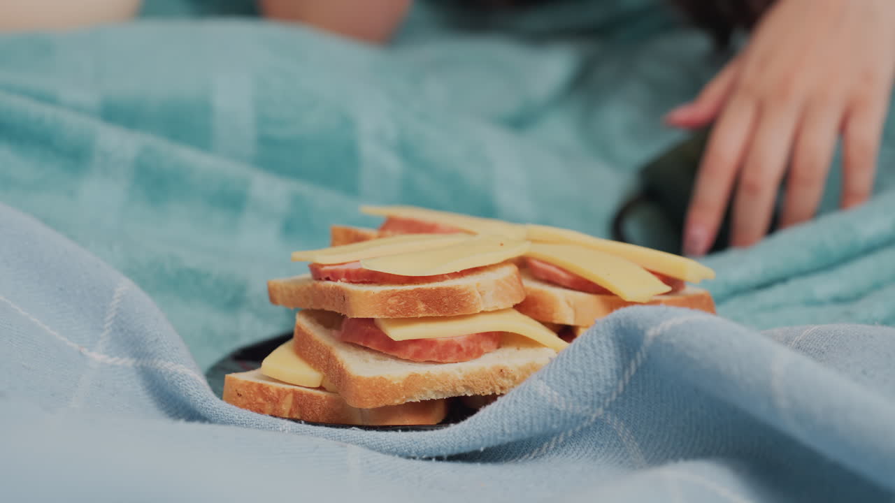 Pile of sliced bread with cheese and meat placed on blue blanket covered bed, with human hand resting close in background, suggesting relaxed indoor meal moment in cozy setting