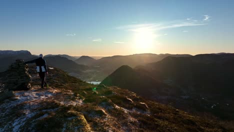 man on mountain peak leaning on stone cairn, watching sunset as aerial moves ahead and passes