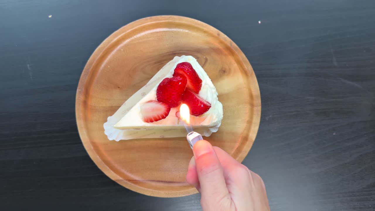 Hand lights small candle on strawberry shortcake slice, overhead view, bright even lighting, static shot