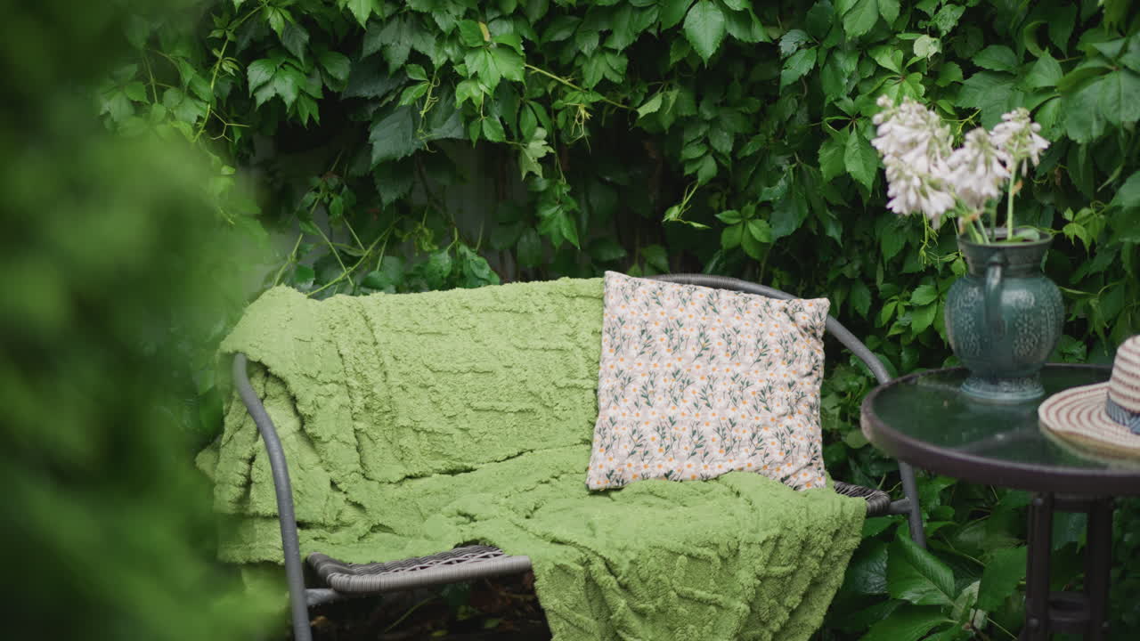 Hyggelig havebænk med grønt tæppe, mønstret pude, rundt metalbord med glasvase med hvide blomster og stråhat, tæt efeu-baggrund, skyggefuld, fredfyldt atmosfære til læsning eller te, morgen.