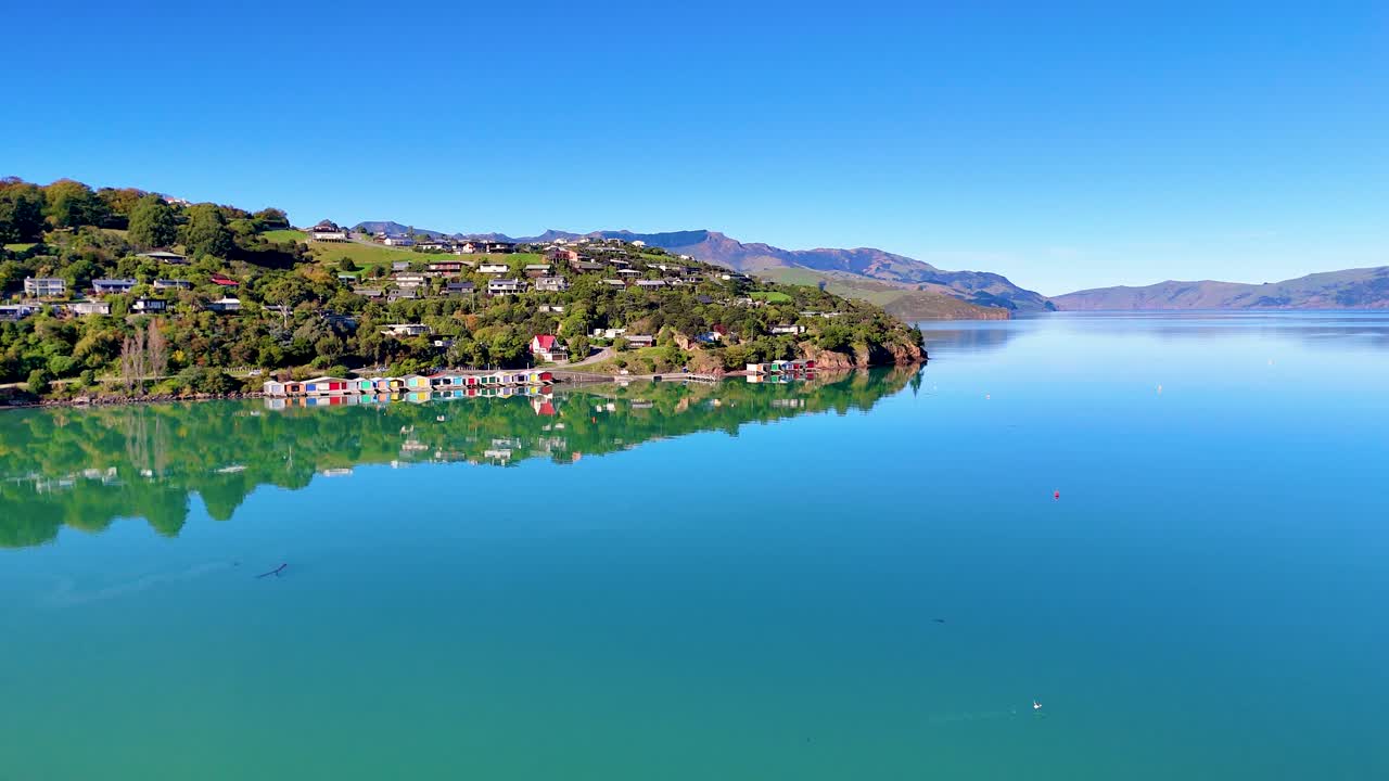 Aerial footage captures Akaroa Harbour's serene waters and lush hills under clear blue skies, showcasing natural beauty and tranquility