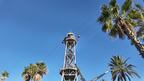 a low angle view of a cable car and tower between palm trees in barcelona city, under a blue sky. timelapse.