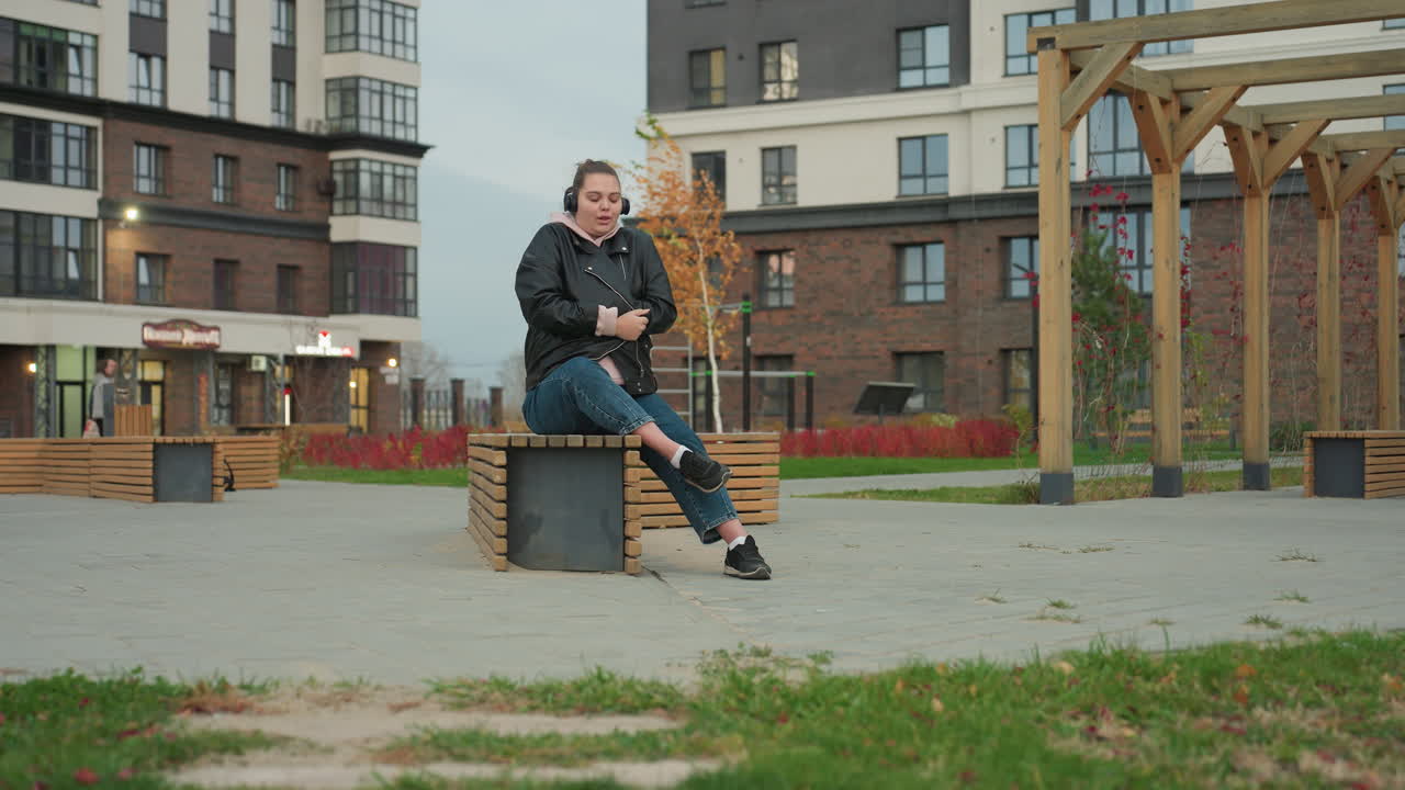White woman seated on wooden bench outdoors adjusts jacket to stay warm while wearing headphones listening to music cool breeze sways nearby plants person walks in distance