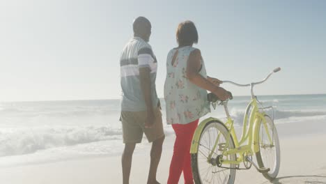 senior african american couple walking with bicycle on sunny beach