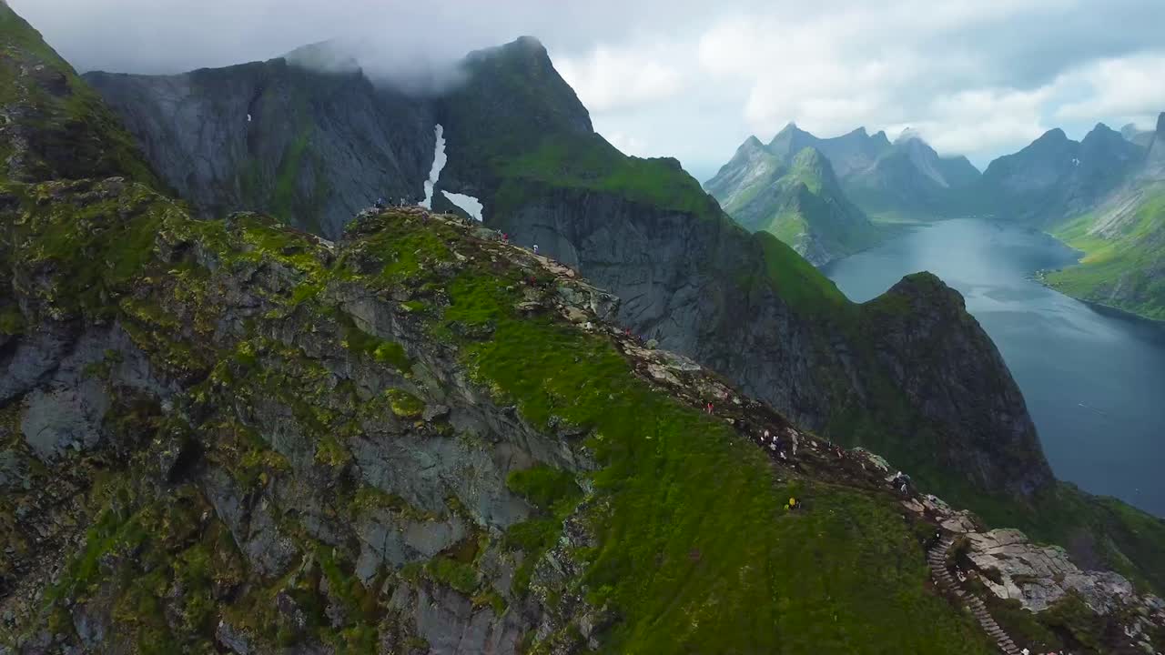 Aerial drone flying closer to a large steep and rocky moss covered cliff or a mountain where tourist and hikers and climbing and walking on the edge in Lofoten Norway. large mountains, lake, clouds.