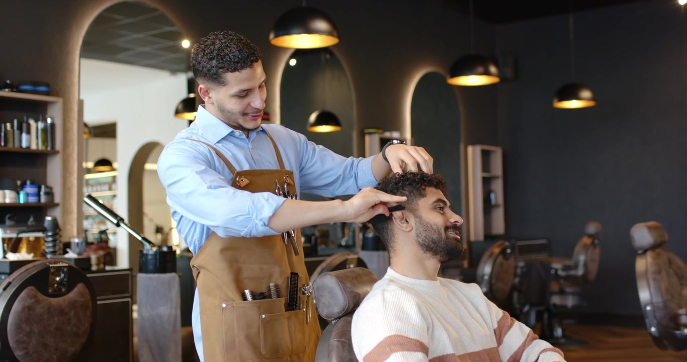 Afroamerikansk barber i læderforklæde klipper siddende kunde med kam og saks i barbersalon.