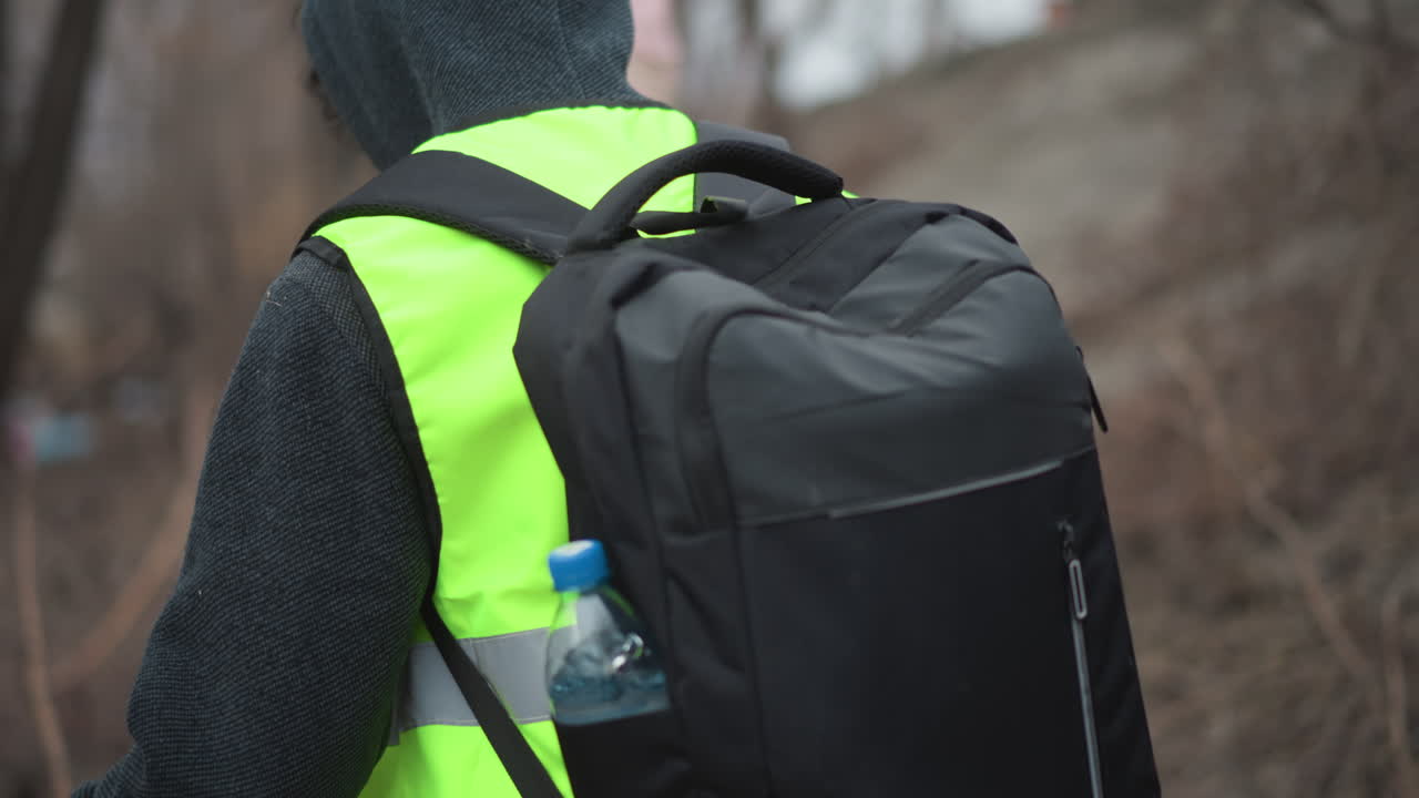 Volunteers in reflective safety vests walking through riverside woodland with backpacks during environmental cleanup, moving carefully among bare branches to collect litter and protect nature