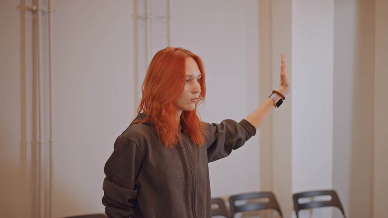 Insegnante di canto dai capelli rossi dirige una prova di scuola di musica in un piccolo studio, gesticolando verso un coro immaginario, sedie pieghevoli vuote, luce calda dalla finestra, espressione concentrata, esercizi di respirazione e tempismo.