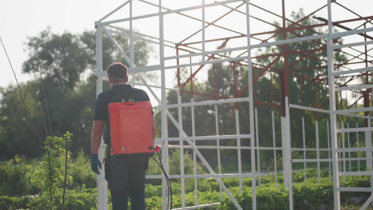 man gardener carrying backpack sprayer wearing mask and sunglasses walking through field with blurred trees background in soft sunlight rural agriculture scene