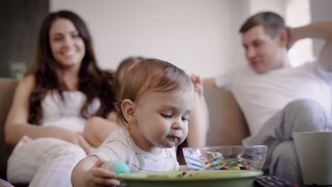 niño pequeño curioso está viendo objetos en la mesa ocasional en la sala de estar del apartamento durante el día, sus padres y hermana mayor en el fondo