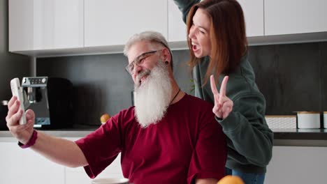 A-happy-elderly-man-with-gray-hair-and-a-lush-beard-in-a-red-T-shirt-takes-a-selfie-using-a-White-phone-with-his-adult-brunette-daughter-in-a-green-sweater-in-a-modern-apartment