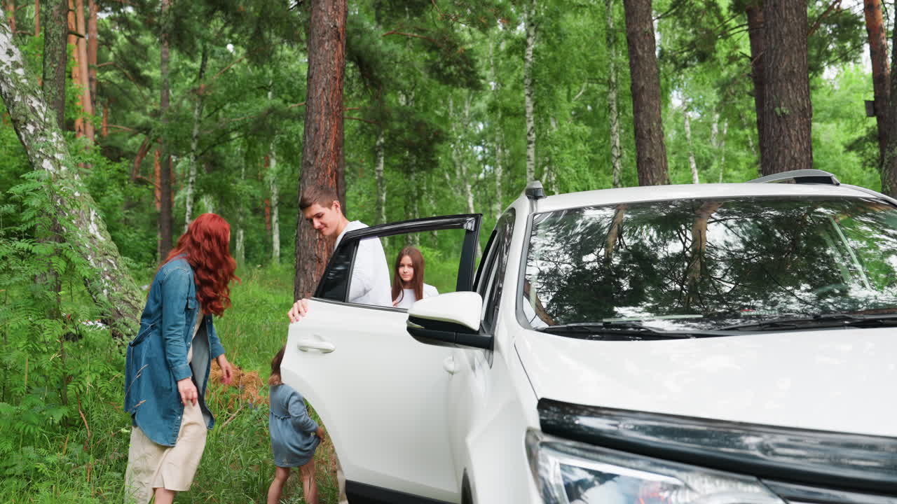 Eldest child carefully carries youngest from back seat of family car parked in forest while mother stands nearby smiling warmly, trees and greenery around, peaceful family bonding moment