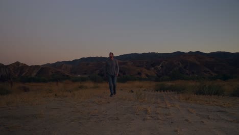 Static-slow-motion-of-a-man-walking-towards-the-camera,-countryside-at-dusk,-sunny-evening-with-mountains-in-the-distance