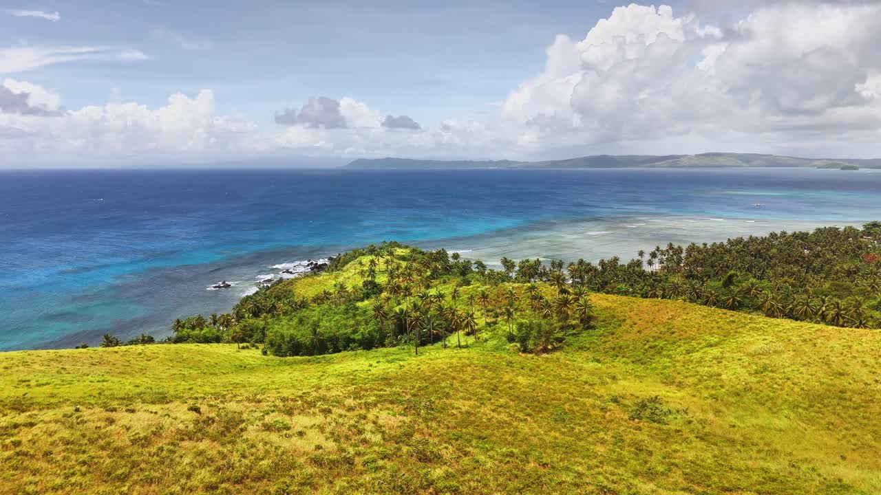 Colinas gramadas se estendem em direção à costa, onde densas palmeiras margeiam as águas azuis claras do Pacífico na Ilha Corregidor, Ilhas Siargao, Filipinas, sob um céu tropical quente.