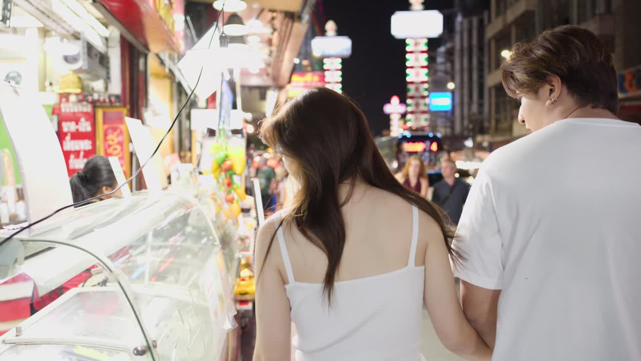 色鮮やかなネオンサインが輝く夜の屋台街を散策する若いカップルの後ろ姿