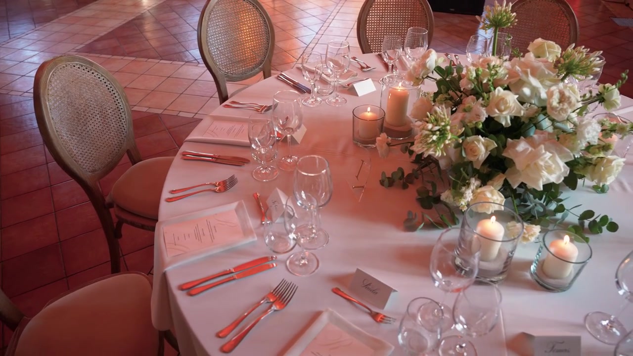 Beautifully arranged round table with white floral centerpiece, glowing candles, fine glassware and menus set for a formal wedding. Warm ambient lighting, sophisticated decor, and elegant atmosphere.