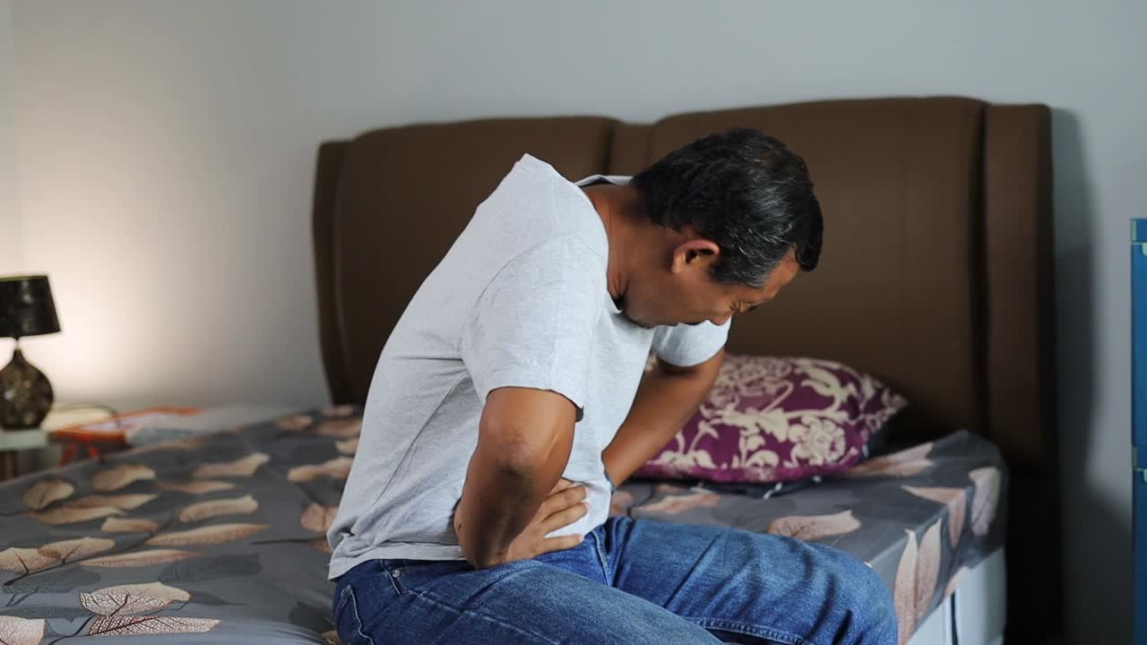 Asian man sitting on bed clutching stomach experiencing intense abdominal pain at home
