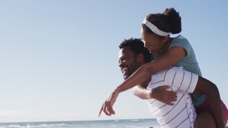 hombre afroamericano sonriendo y llevando a una mujer afroamericana a hombros en la playa