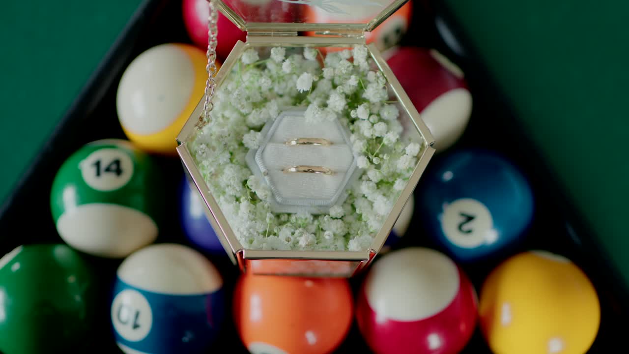 box with two wedding rings inside, surrounded by white flowers, placed on a triangle rack of colorful billiard balls