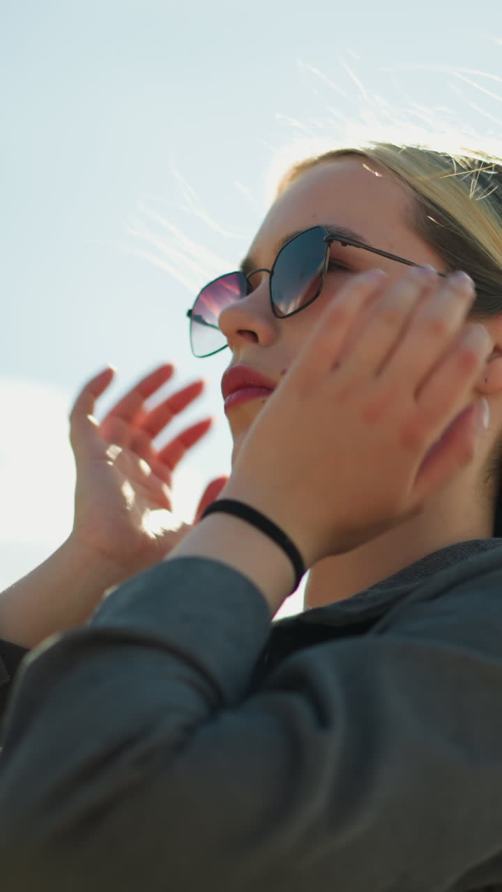 회색 재 아래의 검은 을 입은 여성은 선글라스를 착용하고, 머리카락을 조정하고 집중된 표정으로 목걸이를 만지며, 그녀는 밝은 빛 하늘 아래 야외에 서 있습니다.