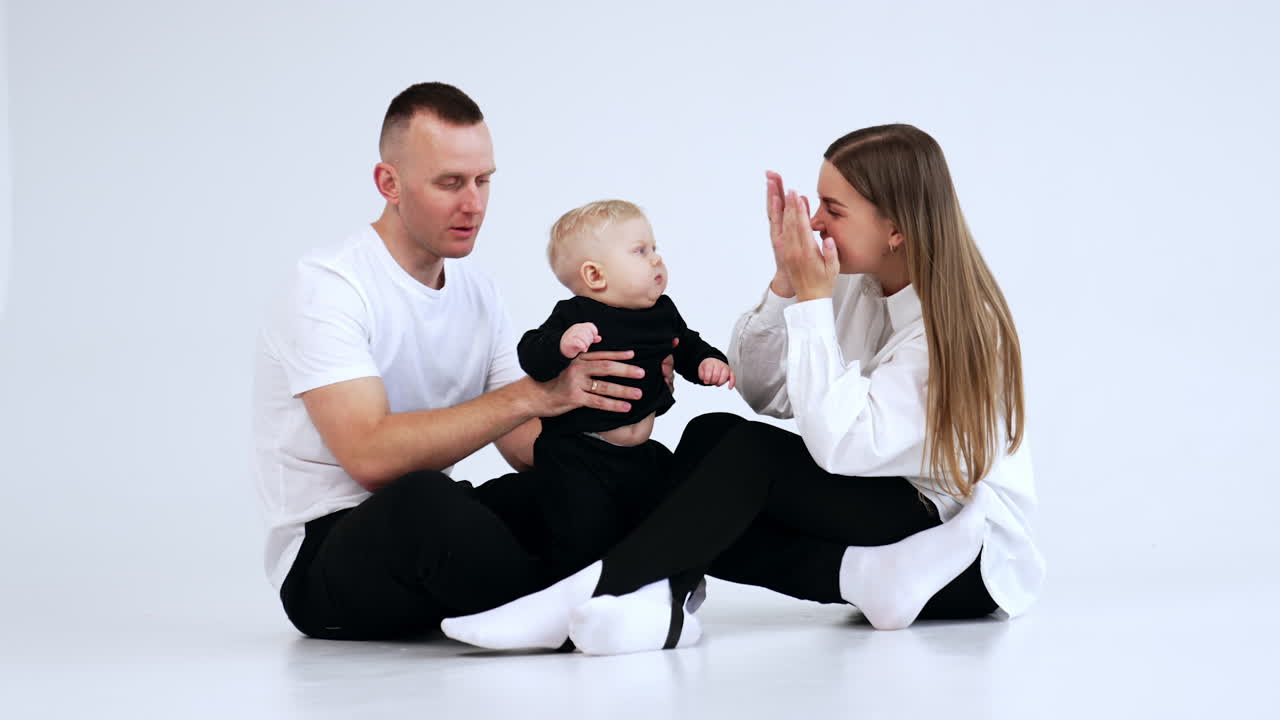 Lovely Caucasian family sitting together in studio. Parents hold their cute little son and mom plays with kid. White backdrop.