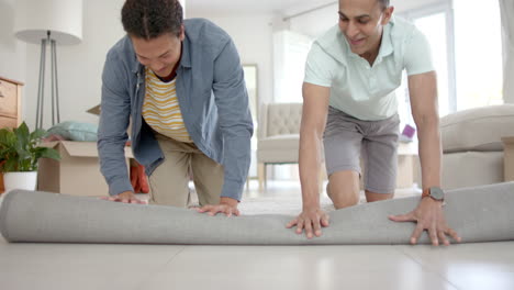 happy diverse gay male couple moving into new home, unrolling rug in living room, slow motion