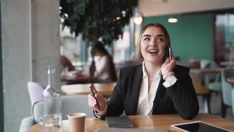 retrato de una joven y hermosa mujer sentada en un café, vestida con ropa de negocios, hablando en el teléfono inteligente con una sonrisa en la cara