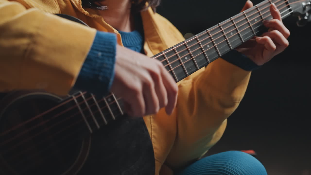 Primo piano mani che strimpellano una chitarra acustica durante una sessione di campeggio con una calda giacca gialla e polsino blu, cantautrice che pratica una calda melodia folk, illuminazione ambientale soffusa, atmosfera intima, attenzione ai dettagli