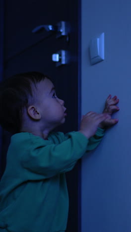 caucasian child stands in the dark room. baby pushes the switch to turn the light on. vertical video