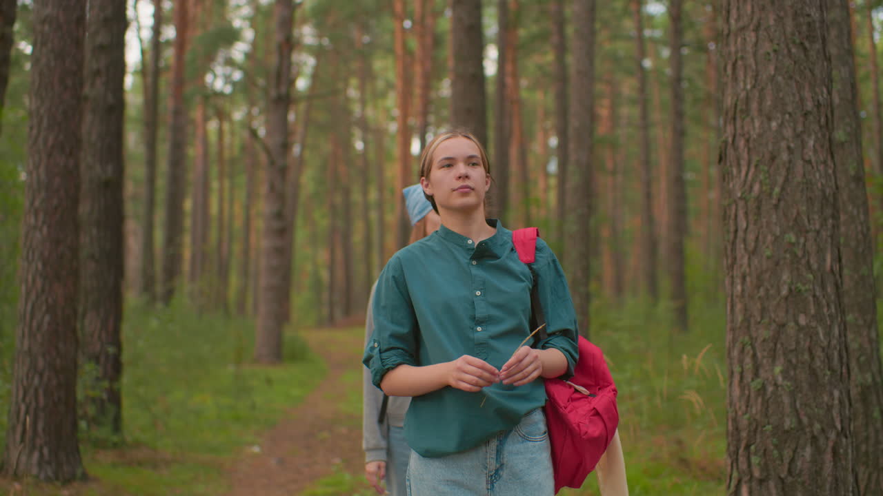 Young woman with a red backpack and green shirt walks through a peaceful forest trail surrounded by tall trees holding a strand of grass with her friend behind her with blue scarf looking down