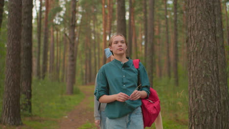 young woman with a red backpack and green shirt walks through a peaceful forest trail surrounded by tall trees holding a strand of grass with her friend behind her with blue scarf looking down