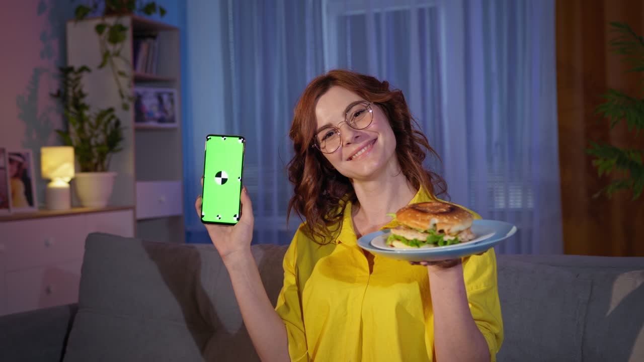 ordenando comida rápida en línea, mujer alegre en vasos con hamburguesa y teléfono móvil con llave de croma verde en las manos sonríe y mira a la cámara durante un bocadillo nocturno en casa