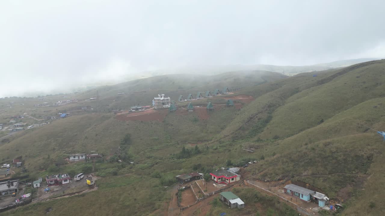 Filmisk luftfoto af det tågede Meghalaya-landskab med små landhuse spredt ud over bølgende grønne bakker under en overskyet himmel.