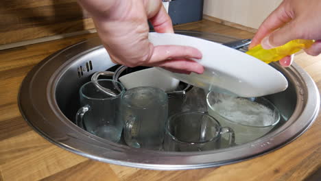 person turns on water and starts washing full sink of dirty dishes, side static view