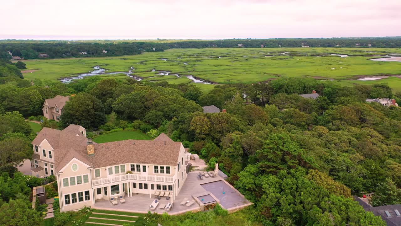 Drone video captures seaside homes perched above sandy bluffs, with stone seawalls lining the beach and lush greenery extending inland toward sprawling marshes in the distance