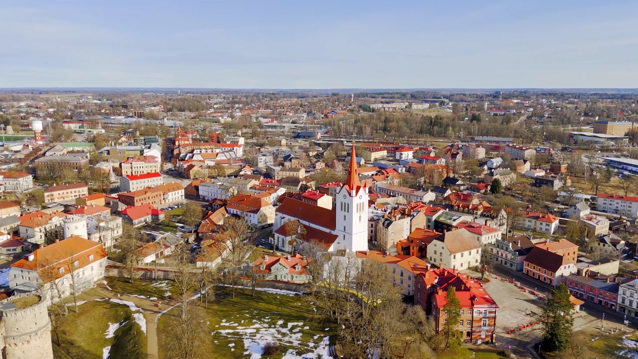 Aerial panorama of Cesis old town with St. John’s Church, castle grounds, red rooftops, and melting snow, capturing historic urban character in early spring light.