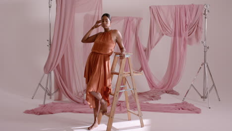 adult indian woman reaching ladder posing in studio, stretching dress and smoothing pink fabric