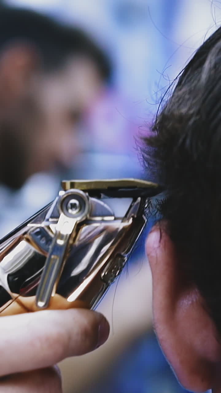 CU shot, slow motion: skilled barber does haircut to young man with black healthy hair using modern shiny golden machine in beauty salon close view