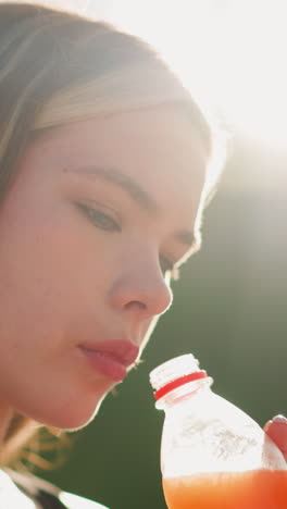 dama bebiendo de una botella de jugo al aire libre, su enfoque atraído a algo ligeramente borroso en la distancia, la luz del sol crea un brillo suave a su alrededor, con un fondo borroso que muestra un poste de lámpara
