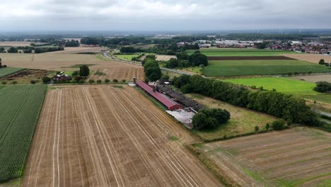 red barn of american farmstead surrounded by agricultural fields on cloudy summer day. rural countryside with small town and wind turbines in distance. aerial orbit wide shot