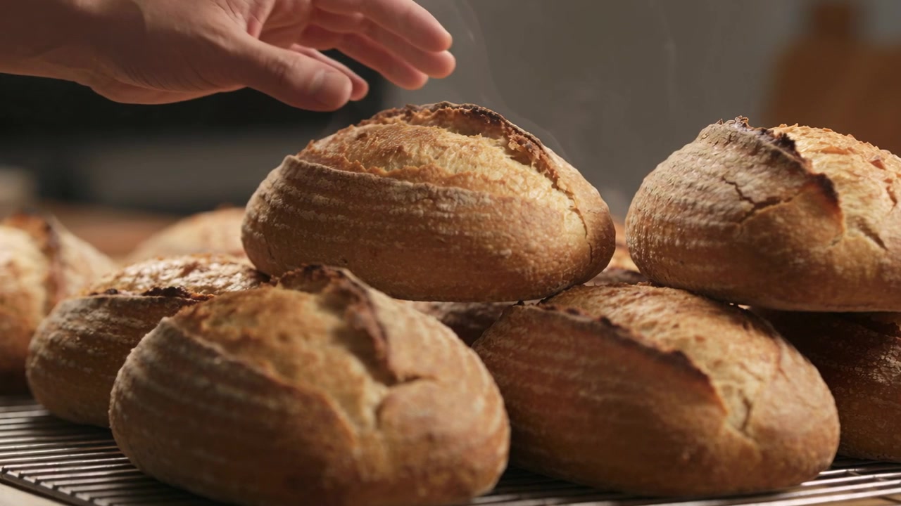 Reaching bare arm, hand entering, cupping, lifting and checking top artisan loaf on metal rack
