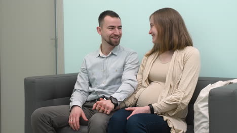 Happy-Pregnant-Couple-Talking-To-Each-Other-And-Holding-Hands-While-Sitting-In-Waiting-Room